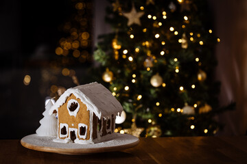 Gingerbread house in living room on a table with Christmas tree in blurry background. Christmas night. Wonderful holiday mood lights. Decorated in festive environment