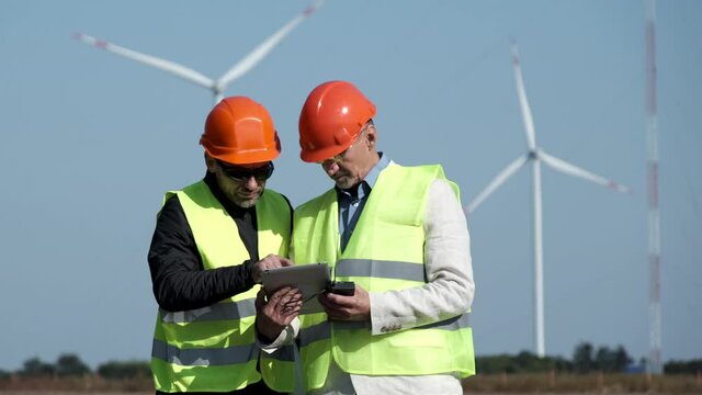 Wind power produced at station under blue sky. Experienced workers control contemporary rotating propellers via tablet at construction site