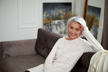 Charming mature female in knitted sweater and with gray hair sitting on comfortable sofa and...