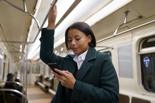 Young afro american woman in metro train using mobile phone, chatting online in social media while returning home after work by public transport. Biracial female suffer from smartphone addiction
