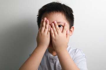 A scared Asian boy covering his eyes with his hands, standing against a white wall.