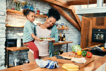 Happy black mother talks on cell phone while preparing breakfast for her daughter in the kitchen.