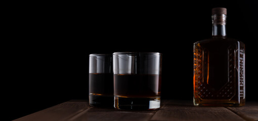 Fototapeta na wymiar Two glasses with alcohol and a bottle on a wooden table in a dark room