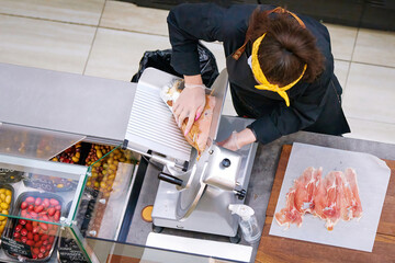 Butcher cut prosciutto, slicing prosciutto on cutting machine. View from above on female butcher...
