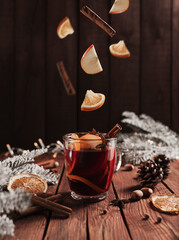 Christmas glass cup or mug with Mulled Wine on wooden table and background with levitating cinnamon stick, dried or sublimated orange, apple slices, present gift boxes, garlands, decoration lights