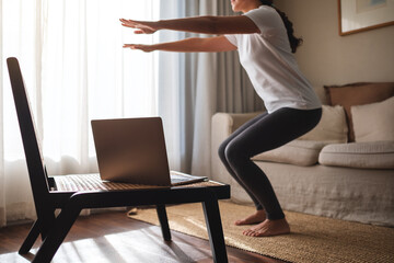 Closeup of a young woman doing squats while watching online workout tutorials on laptop at home