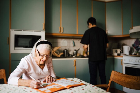 Elderly woman solving crossword puzzle in book while male nurse working at kitchen