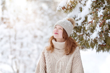 A young woman in a knitted beige hat and sweater admires the winter forest on a sunny day. Winter joys, winter holidays, outdoor recreation concept.
