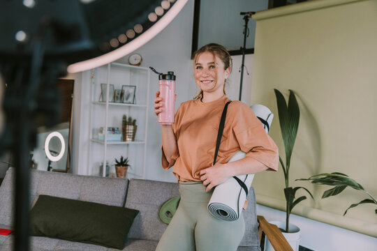 Smiling female influencer with exercise mat and smoothie vlogging at home - Powered by Adobe