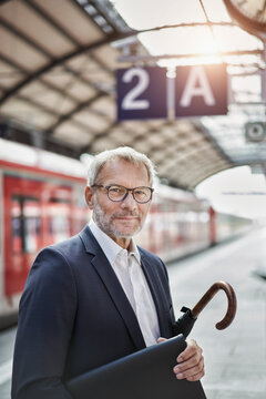 Senior businessman with laptop and umbrella standing on railroad platform