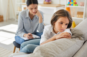 Frustrated little child at psychologist's office. Portrait of girl with sad face sitting on couch during therapy session. Therapist trying to help unhappy, resentful kid who has behaviour issues - obrazy, fototapety, plakaty