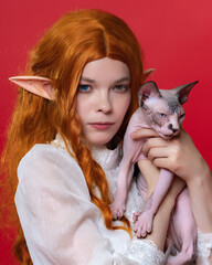 Portrait of cosplayer elf young female in white dress, with beautiful red long hair, eyes of different colors who looks at camera thoughtfully, holding Sphinx kitten in her hands. Red background.