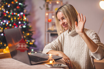 Excited adult 40s woman waving hello with hand, using laptop at home