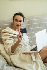 young girl in home clothes with a card in her hands and a laptop on her knees shopping online.