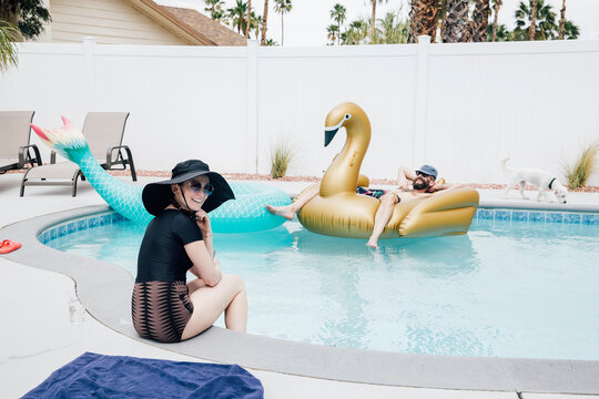 Smiling woman sitting at poolside while man relaxing on Inflatable swan during sunny day