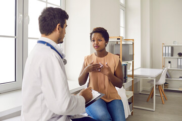 Black woman visiting hospital. Female patient sitting on couch in exam room and talking to professional doctor about chest pain. General practitioner listening to unhappy young lady asking for help - Powered by Adobe
