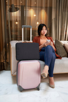Young woman with mobile phone sitting on sofa by luggage in hotel
