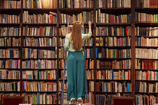 Young woman standing on ladder while searching book in library