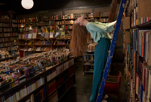 Redhead woman with head back standing on ladder in library