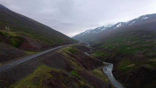 Aerial flying through a valley in Iceland with river and road below. Cars driving along the road, surrounded by hills and snowy mountains. Cloudy overcast day.