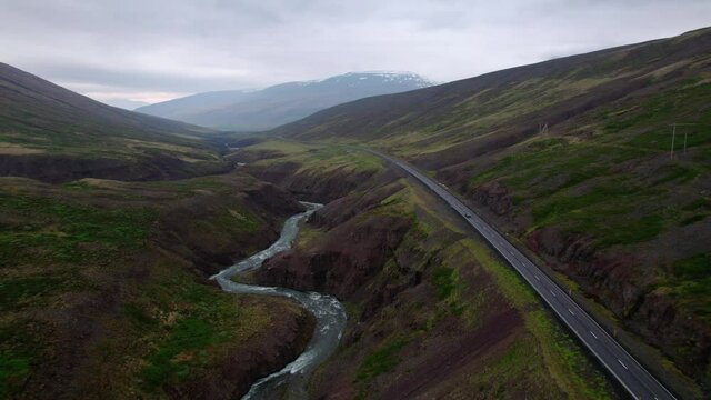 Backwards flying aerial shot in dramatic Iceland landscape with car driving on road below. Dark cloudy day, with river below.