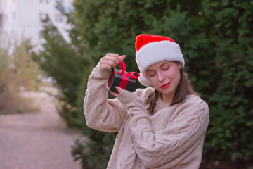 Close up woman in Christmas cap shaking red box at her ear trying to guess what is inside standing