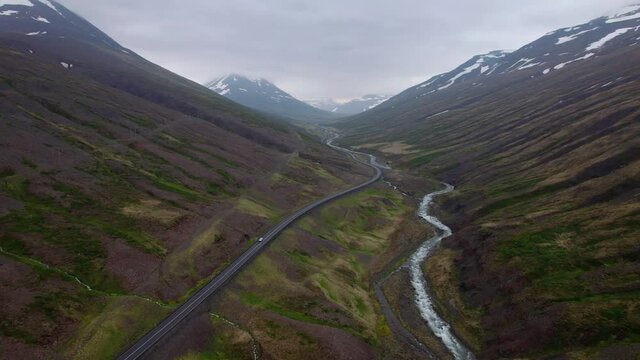 Turning aerial shot with wide view of Icelandic valley and mountains. Car drives below near a river. Dramatic landscape.