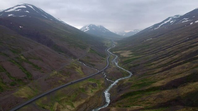 High aerial shot over Icelandic landscape of valley and mountains. Rivers wind through and cars drive below. Overcast day.