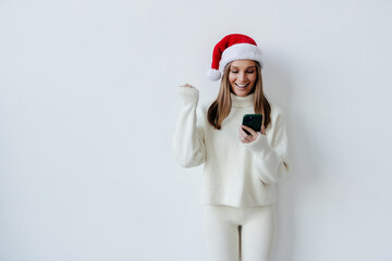 Excited young Santa woman in Christmas hat hold use mobile cell phone type say yes found out big win doing winner gesture isolated on white background