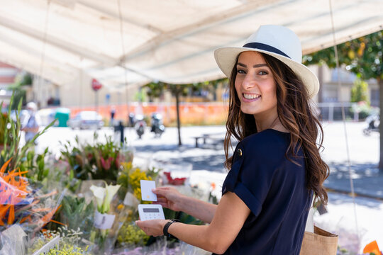 Smiling young woman in sun hat paying with credit card at florist shop