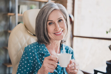 Photo of cheerful aged woman happy positive smile relax break pause drink hot coffee tea enjoy indoors