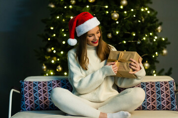 Winter, happiness, holidays and people concept. smiling woman in santa helper hat with gift box over living room and christmas tree background