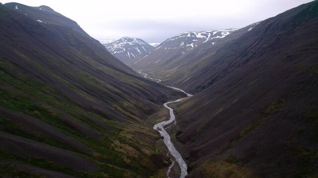 Aerial flying over dramatic Iceland landscape in valley with mountains in distance. Dark ground and river below.