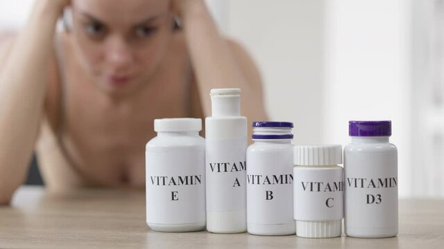 Row of vitamin bottles on table with blurred Caucasian young woman at background thinking. Pharmaceutical nutritional supplements indoors with slim beautiful millennial lady choosing pills