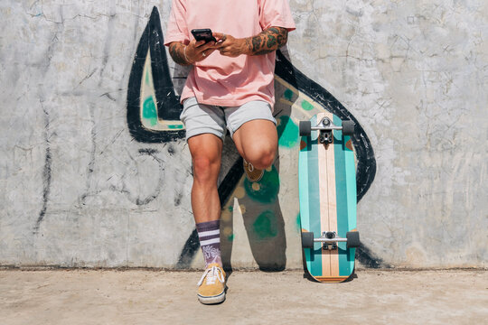 Man text messaging through smart phone by skateboard while leaning on wall during sunny day
