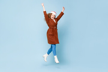 Fototapeta na wymiar Pretty Caucasian woman wearing Christmas hat and coat jumping with hands raising in isolated light blue color background