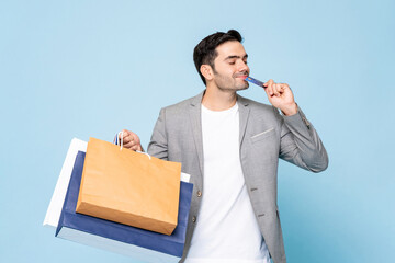 Young happy handsome Caucasian man holding shopping bag and credit card in isolated light blue background - obrazy, fototapety, plakaty