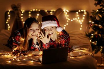 Two charming sisters lie on the bed with a laptop on Christmas or New Year's eve and congratulate their friends and relatives online. Christmas greetings online, virtual party for Christmas