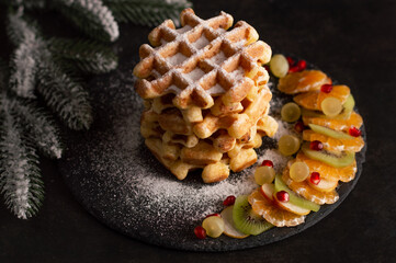 Belgian waffles with powdered sugar and slices of fruit on a dark background with sprigs of spruce.