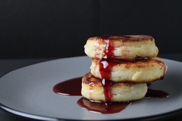 food breakfast cheesecakes curd pancakes three pieces lie stack of slides on top of each other and pour jam sauce cherry red on a gray plate on a black background
