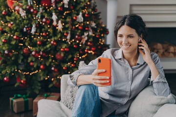 Happy hispanic woman in wireless earphones holding phone, sitting near Christmas tree at home