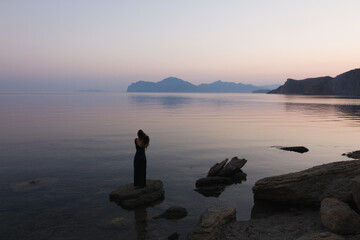 Silhouette of a girl by the sea with stones in the foreground and rocks in the background against the background of a calm sea dreaming suffering enamored inspired