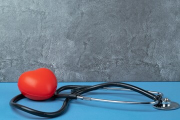 A stethoscope and a rubber red heart on a blue table against a gray marble wall. Heart health, healthcare concept.