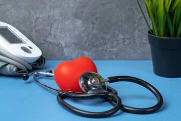 Stethoscope, blood pressure monitor and rubber heart on a blue table with a plant against a gray marble wall. Heart health, healthcare concept.