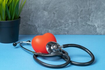 Stethoscope and rubber red heart on a blue table with a plant against a gray marble wall. Heart health, healthcare concept.