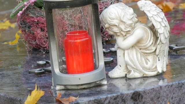 Red memorial candle burning with fire on rainy day on tombstone in cemetery autumn, rainy day. Grave with tombstone on raindrops falling, against abackground of yellow foliage next to sad angel statue