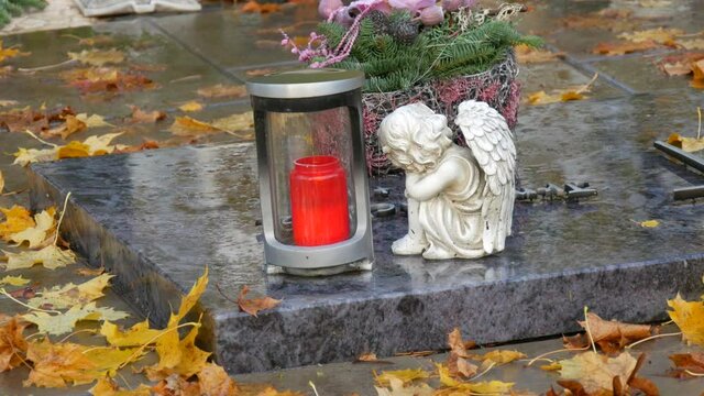 Red memorial candle burning with fire on rainy day on tombstone in cemetery autumn, rainy day. Grave with tombstone on raindrops falling, against abackground of yellow foliage next to sad angel statue
