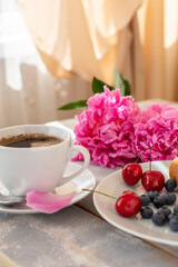 Fototapeta na wymiar Morning coffee, blueberries and cherries, beautiful pink peony flowers on the table in the rays of the sun from the window. Cozy romantic breakfast