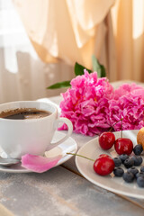 Morning coffee, blueberries and cherries, beautiful pink peony flowers on the table in the rays of the sun from the window. Cozy romantic breakfast