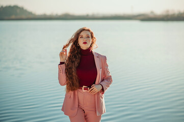 Fashionable freckled readhead woman with natural long curly hair, wearing burgundy color cashmere turtleneck sweater, pink suit with  blazer, belt, trousers.  Copy, empty space for text